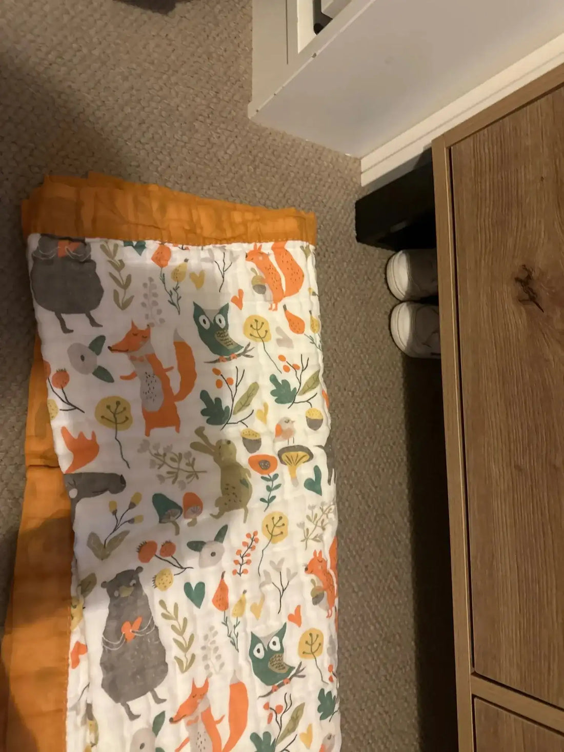 Une couverture douillette en tissu blanc avec un imprimé animalier ludique de forêt mettant en scène des renards, des hiboux et des lapins aux tons orange, vert et gris, bordée d'un liseré orange.